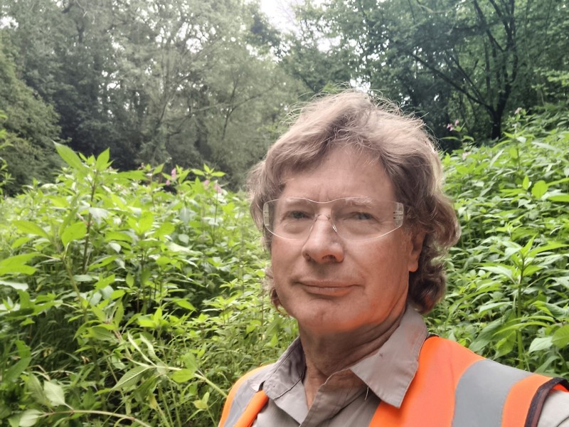 A man in an orange high-vis vest and safety goggles stands infront of a dense stand of plants that are as tall as him