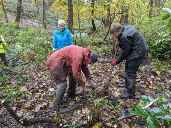 three people wearing waterproof clothing are digging in soil in a woodland